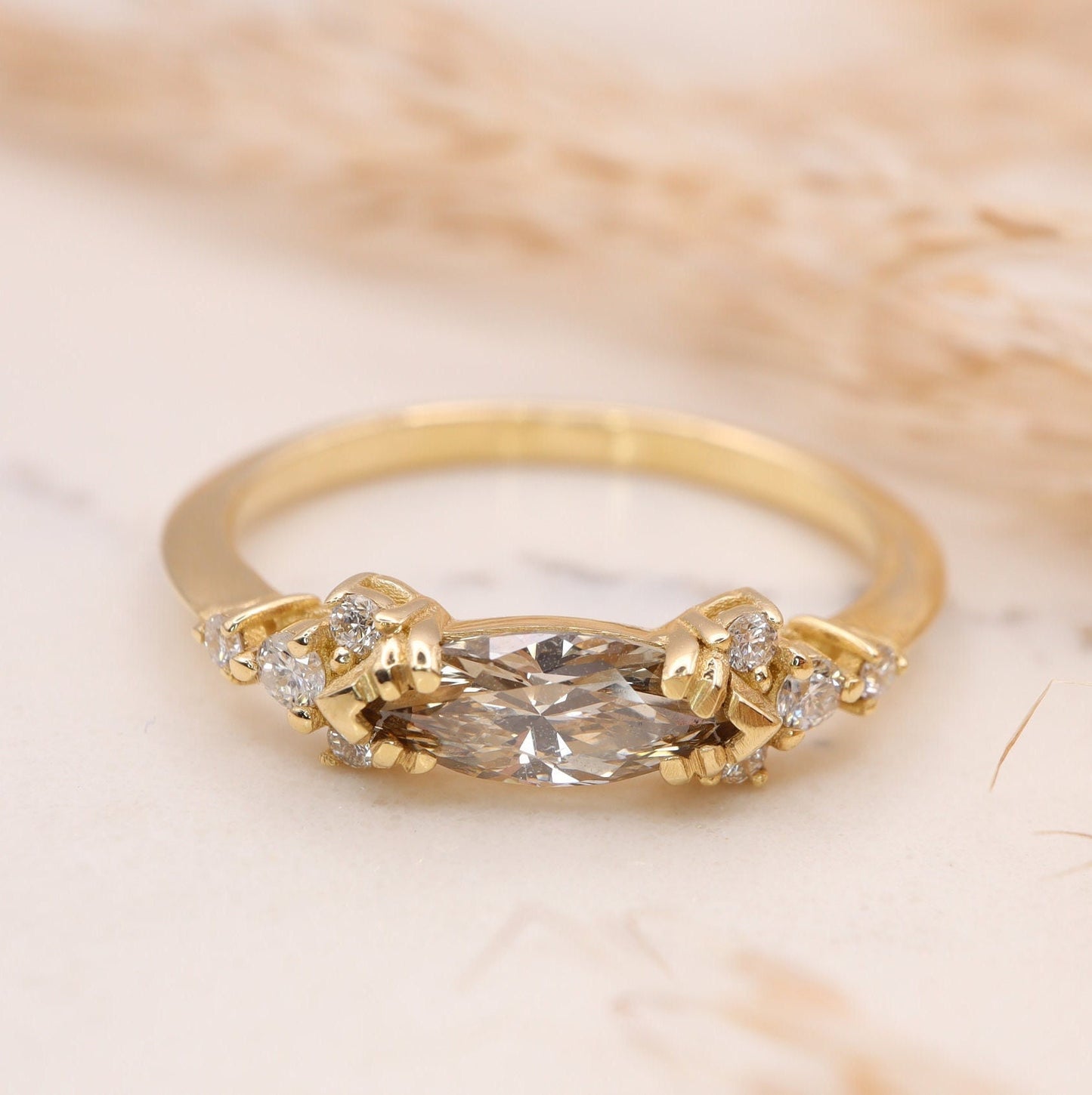 A champagne marquise-cut diamond engagement ring on a marble slab, facing forward, featuring four small diamonds on each side, inspired by Art Deco