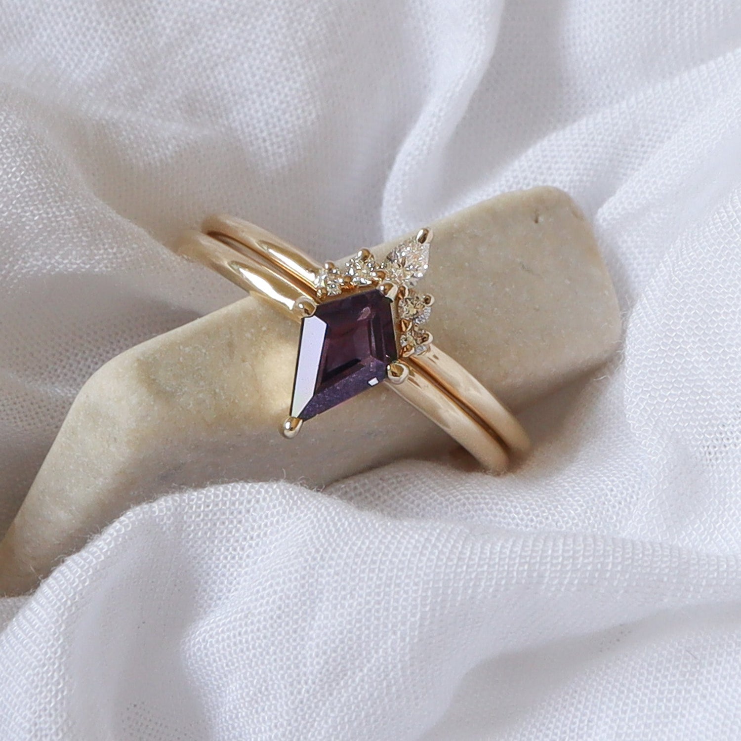 סט טבעות זהב צהוב מבריק, הכולל טבעת עם ספיר סגול טבעי לא מחומם בצורת מגן וטבעת משלימה עם יהלום טיפה ויהלומים עגולים קטנים, מונחות על בד לבן ואבן שיש בהירה.