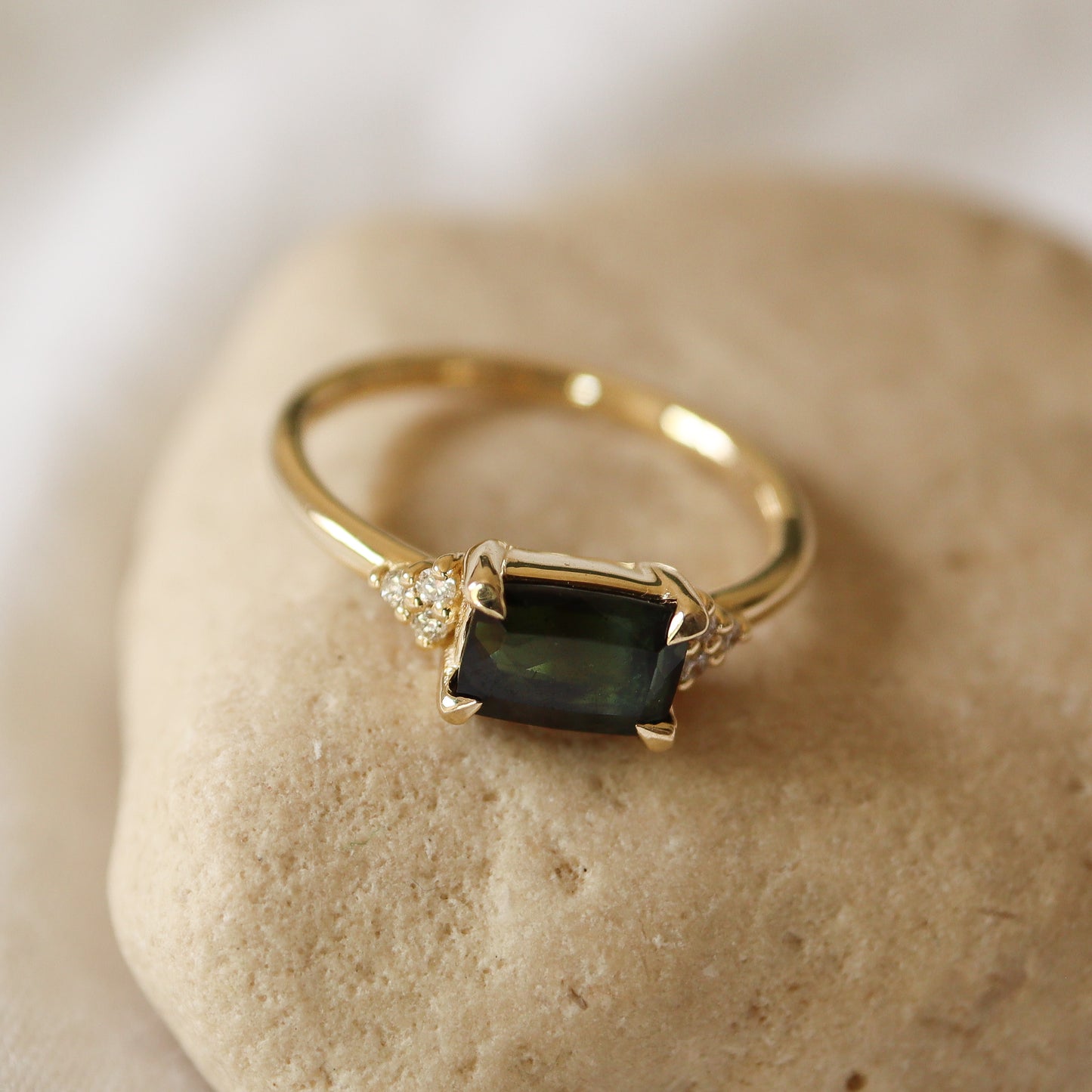 טבעת טורמלין ירוק כהה בזהב צהוב עם שישה יהלומים קטנים מכל צד, מונחת על רקע אבן טבעית.