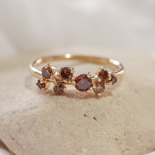 טבעת זהב צהוב עדינה בעיצוב קלאסטר א-סימטרי, משובצת יהלומים טבעיים עגולים בגווני חום, כתום ואדמדם, מונחת על אבן טבעית בהירה.