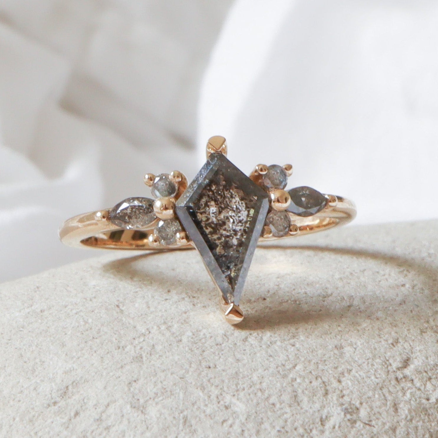 טבעת זהב צהוב עם יהלום מלח-פלפל כהה בצורת דלתון במרכז, ומשני צדדיו יהלומים קטנים בחיתוך מרקיזה ועגול במבנה פרחוני על רקע טבעי.