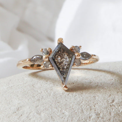 טבעת זהב צהוב עם יהלום מלח-פלפל כהה בצורת דלתון במרכז, ומשני צדדיו יהלומים קטנים בחיתוך מרקיזה ועגול במבנה פרחוני על רקע טבעי.