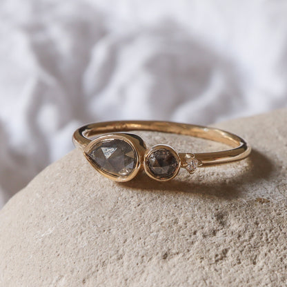 טבעת זהב 14 קראט עם יהלום מלח-פלפל בצורת טיפה ויהלום עגול בשיבוץ כוס מגן על רקע אבן.