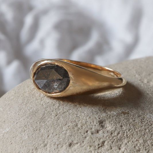 Oval cut salt and pepper diamond in a shiny finish signet ring design resting on a rustic showing its rustic touch.