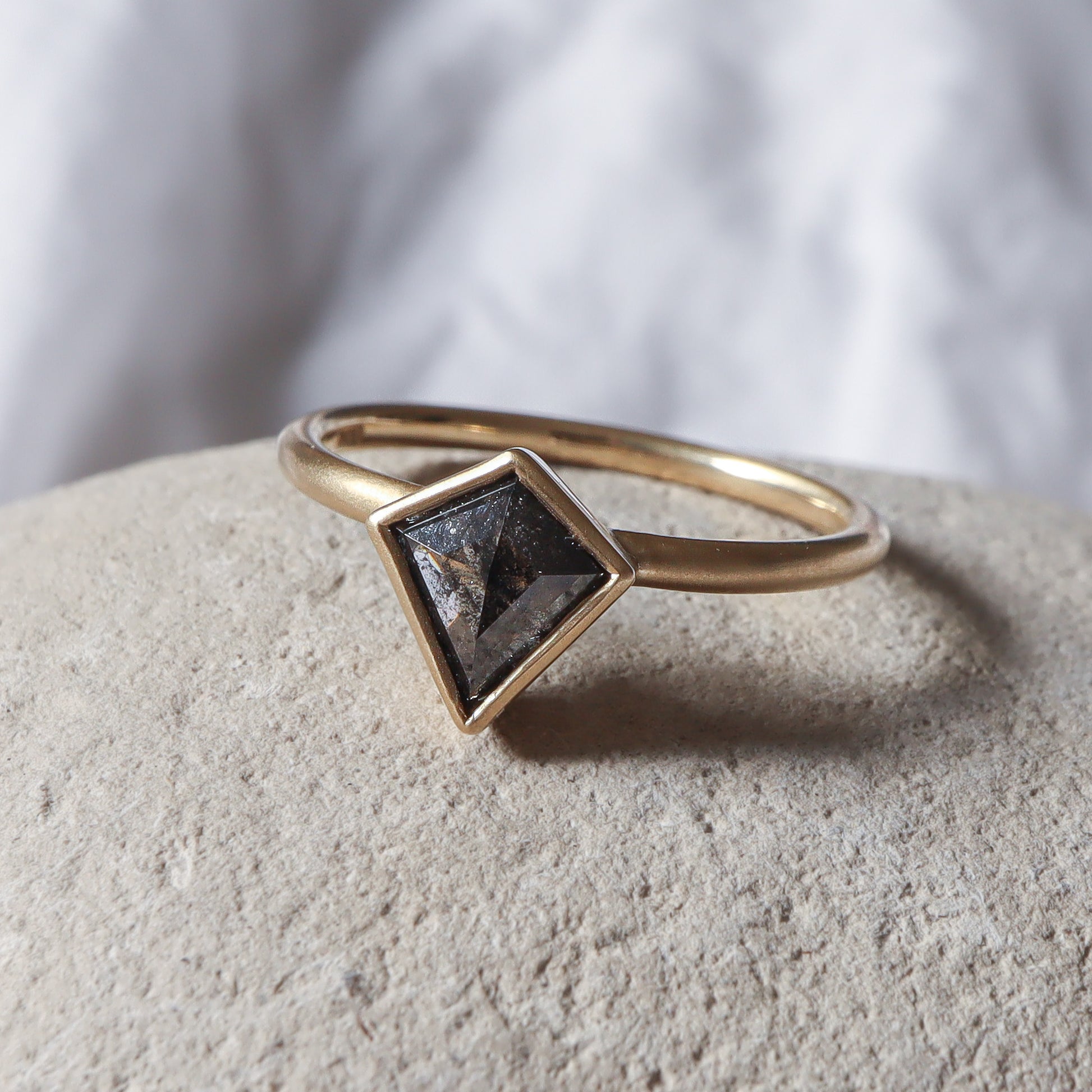 טבעת יהלום משובצת יהלום בצורת דלתון בגוון אפור כהה, הטבעת זהב צהוב עם גימור מט גולמי.