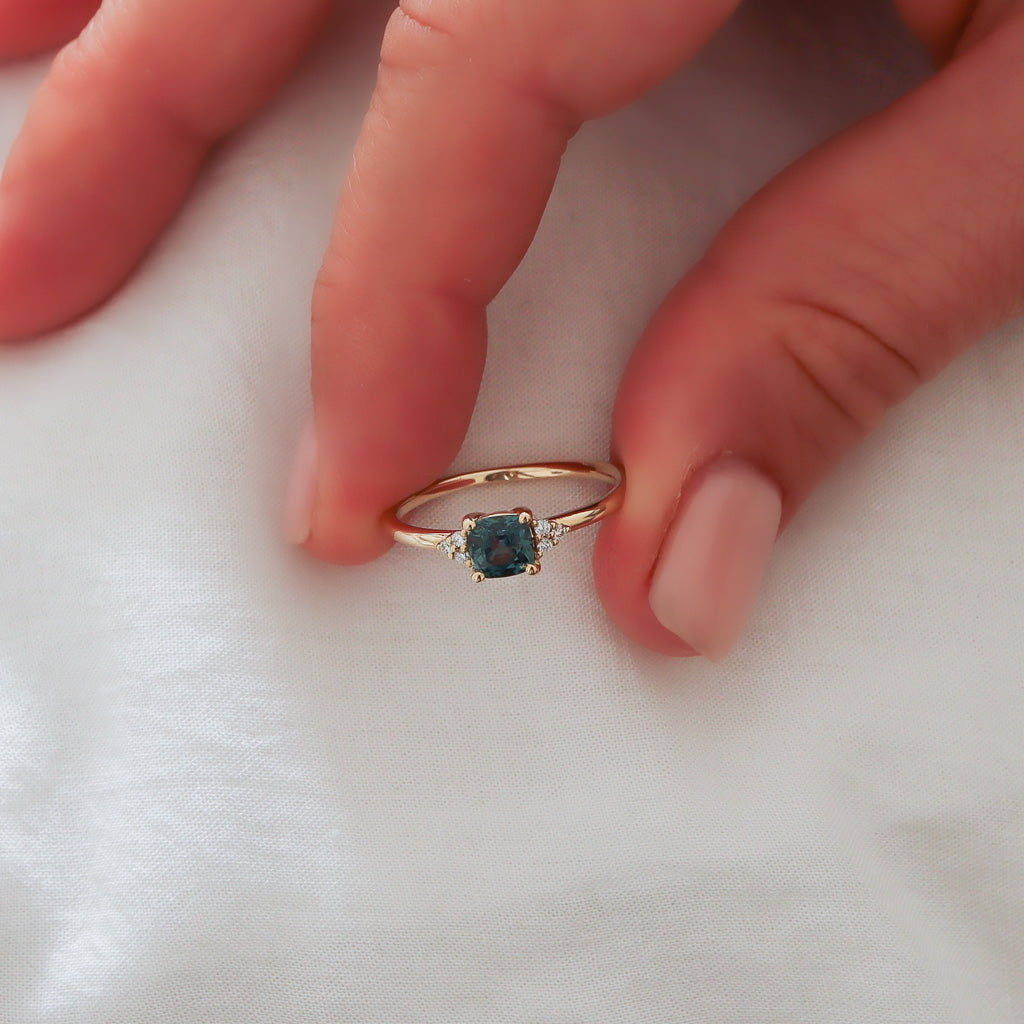 The teal sapphire ring held gently between fingers against a white linen fabric, revealing the brilliance of the diamonds.
