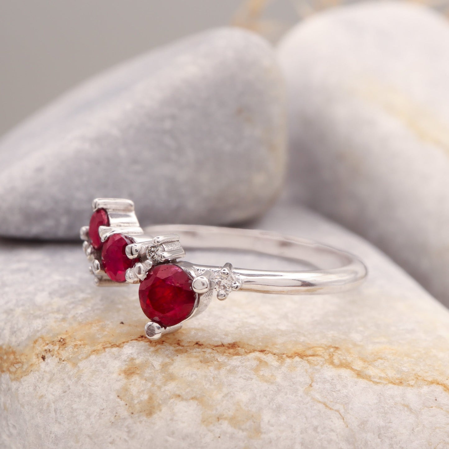 Profile view of a 3 round Ruby and 4 small round Diamond cluster ring in solid white gold, displayed on a grey stone.