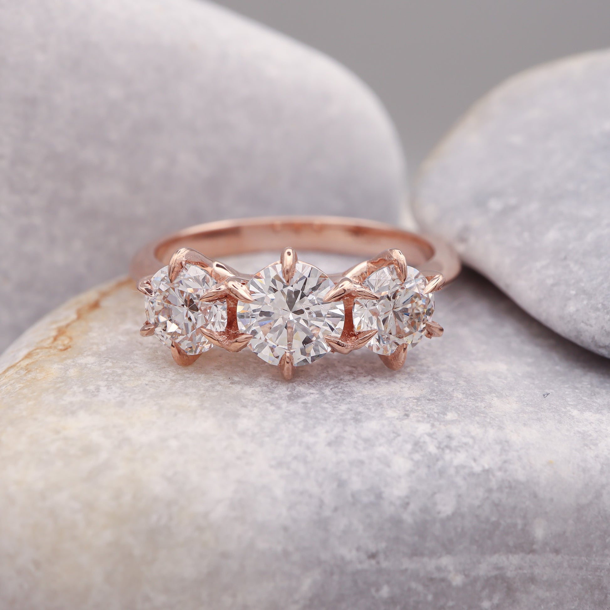 three diamond rose gold ring with delicate 6 prong design, on a grey stone.