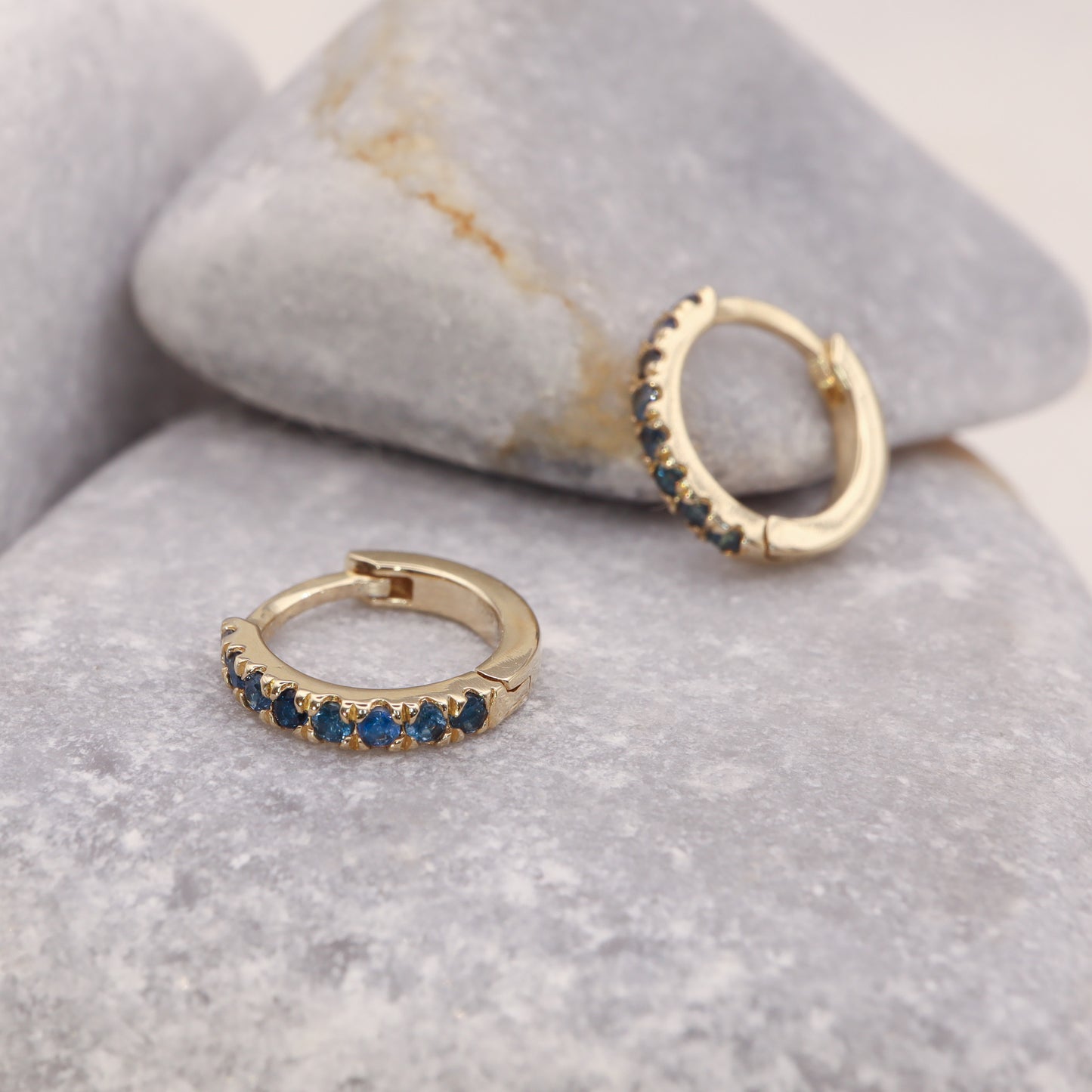 Gold huggie hoop earrings featuring blue sapphires, in a delicate pave setting, displayed on a grey stone.