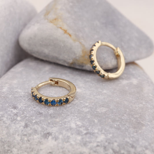 Gold huggie hoop earrings featuring blue sapphires, in a delicate pave setting, displayed on a grey stone.