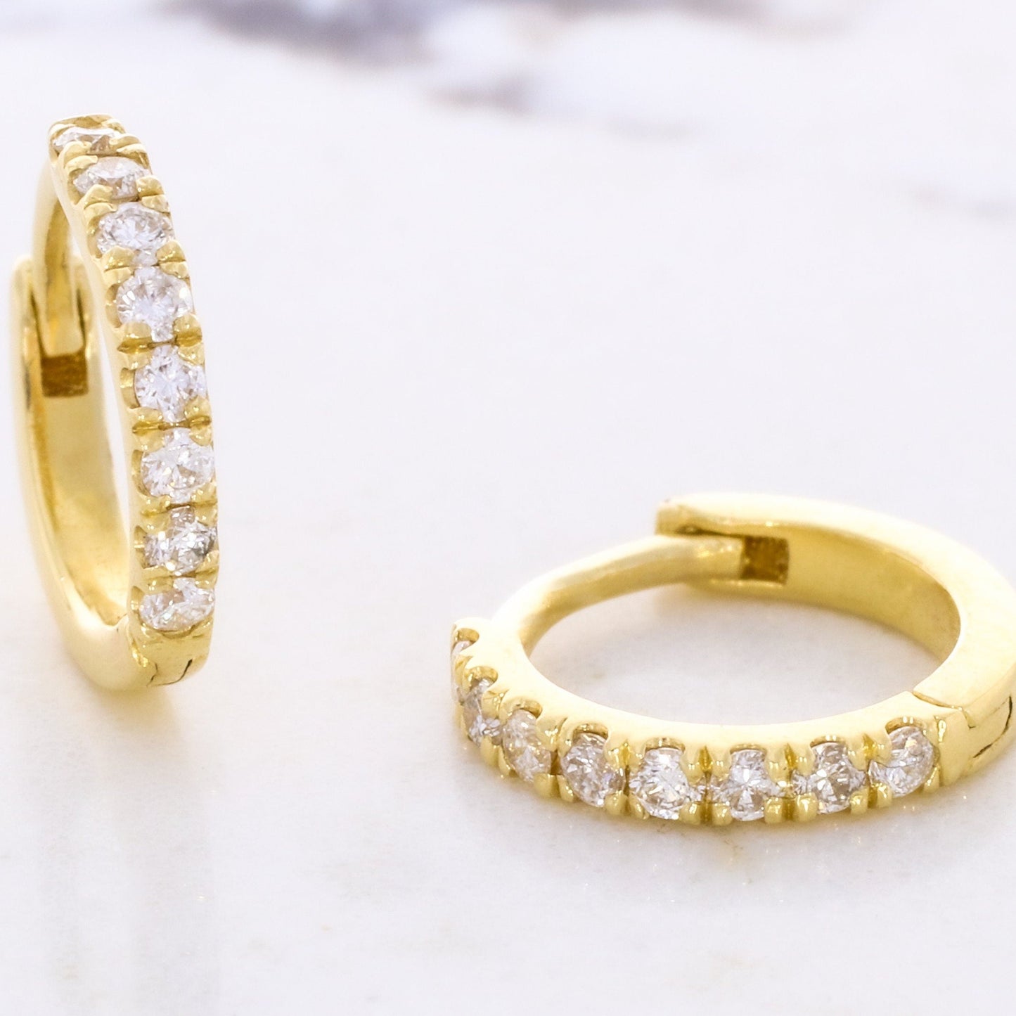 One diamond huggie earring lies flat, while the other stands upright, showcasing their pave setting on a white background