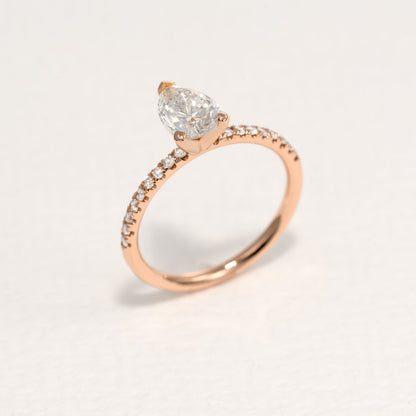 Side view of a pear-shaped diamond set in a two-tone design with round side diamonds, showcased with a rose gold band, on a white background