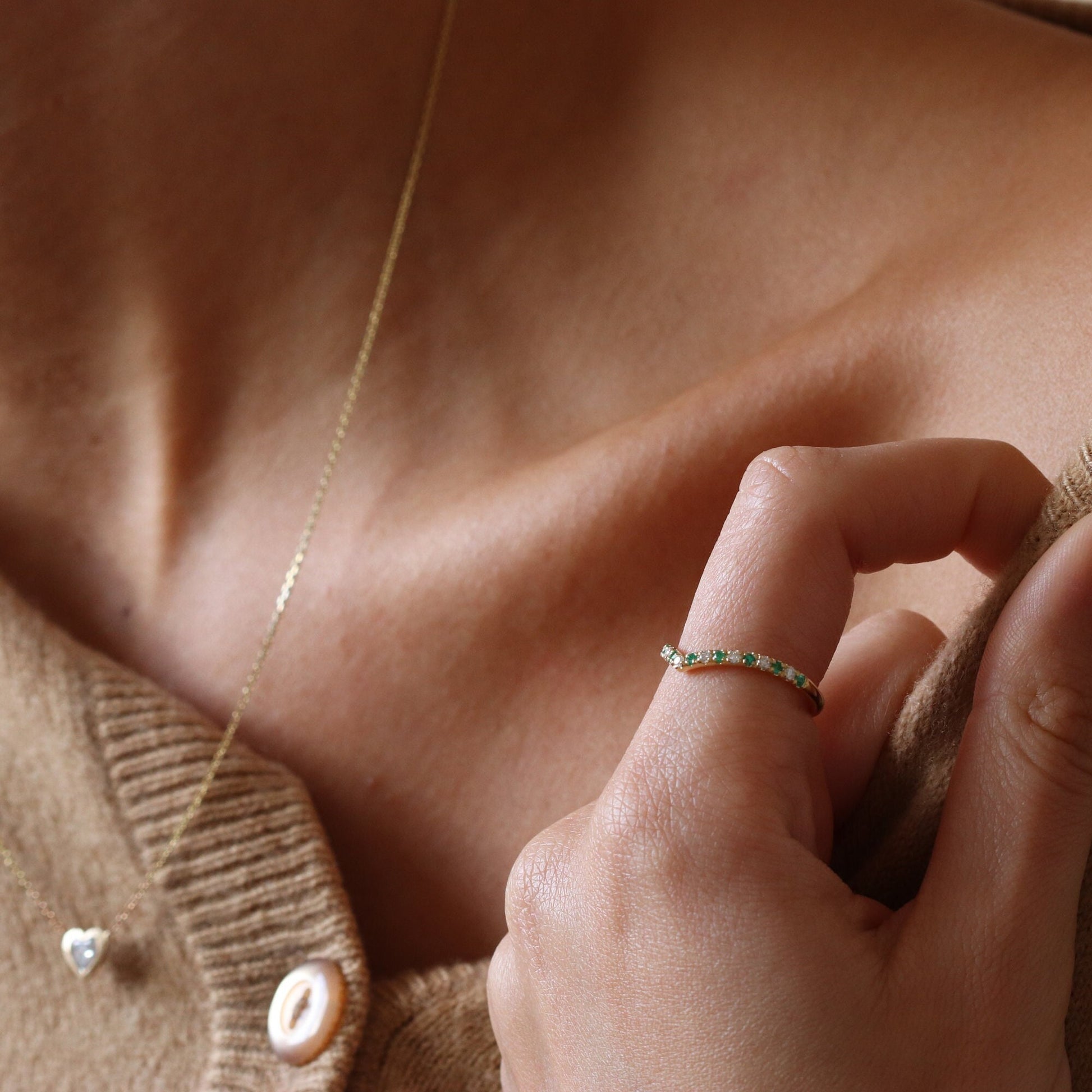 Gold Curved Emerald and Diamond Eternity Stacking Ring, along with a heart-shaped bezel diamond gold necklace, on a woman's hand