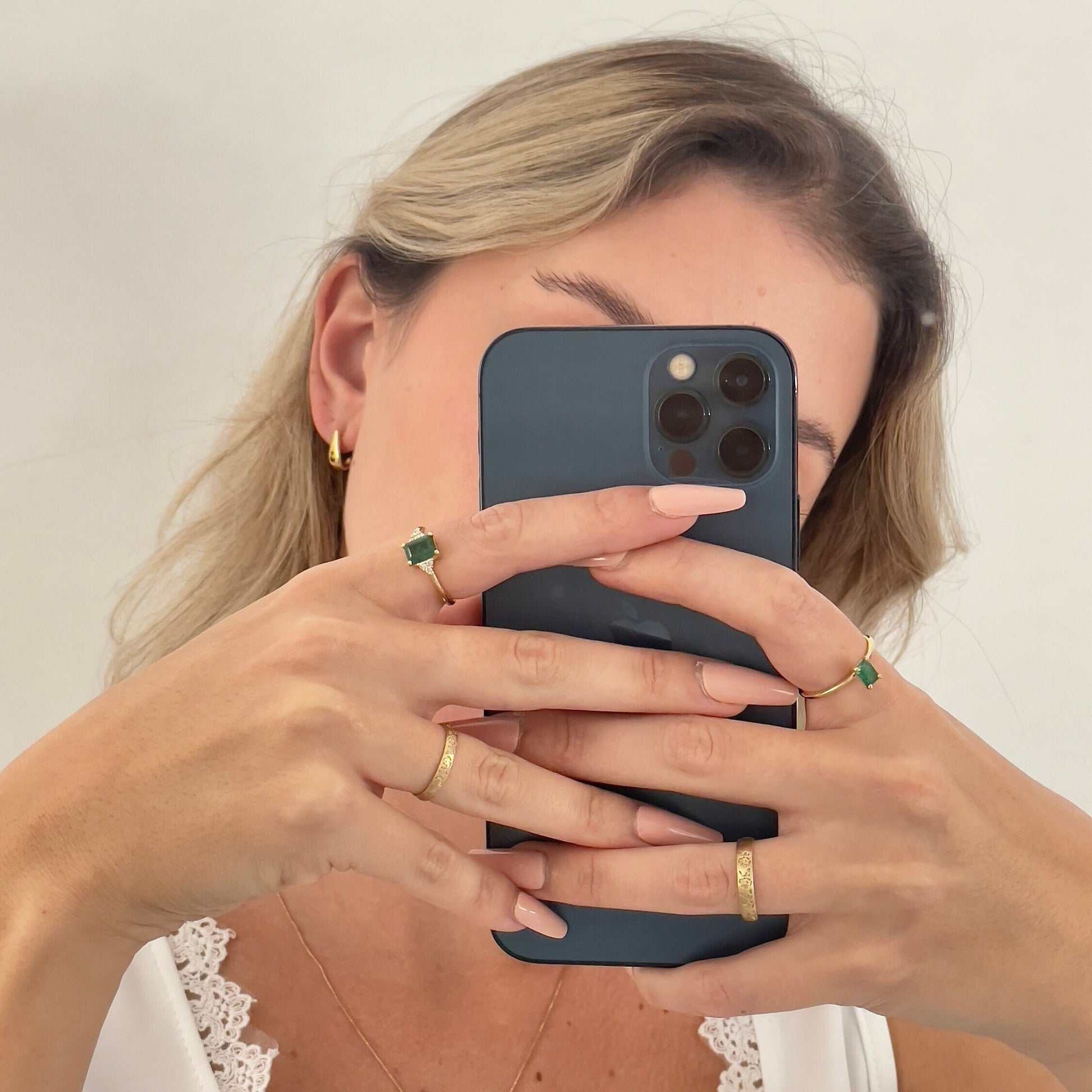 Woman's hand holding a phone in front of her face, wearing a gold solitaire ring with a baguette-shaped emerald, along with two gold wedding bands and another emerald ring with smaller diamonds