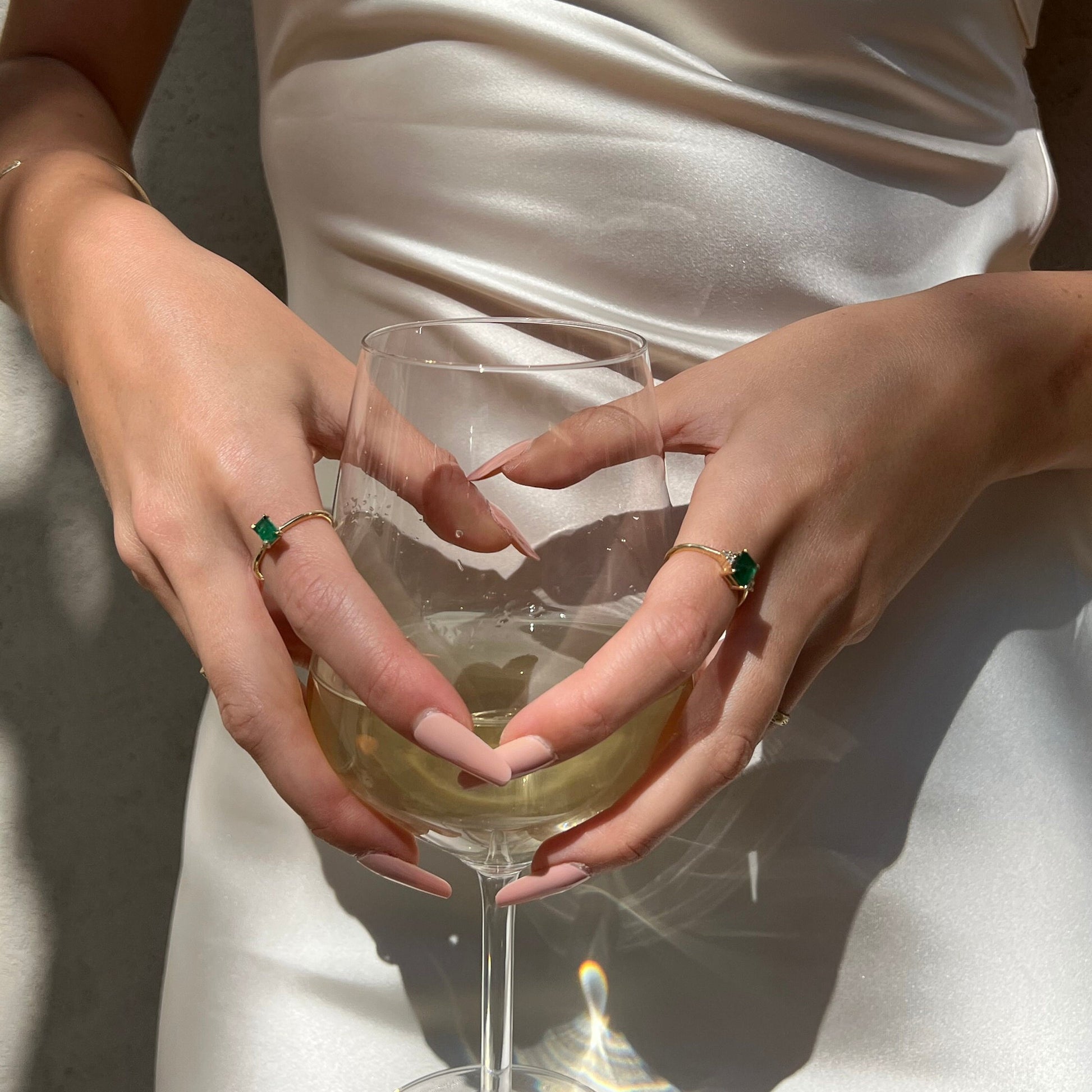 Woman's hand holding a glass of wine, adorned with a gold solitaire ring featuring a baguette-shaped emerald, alongside another emerald ring with smaller diamonds.