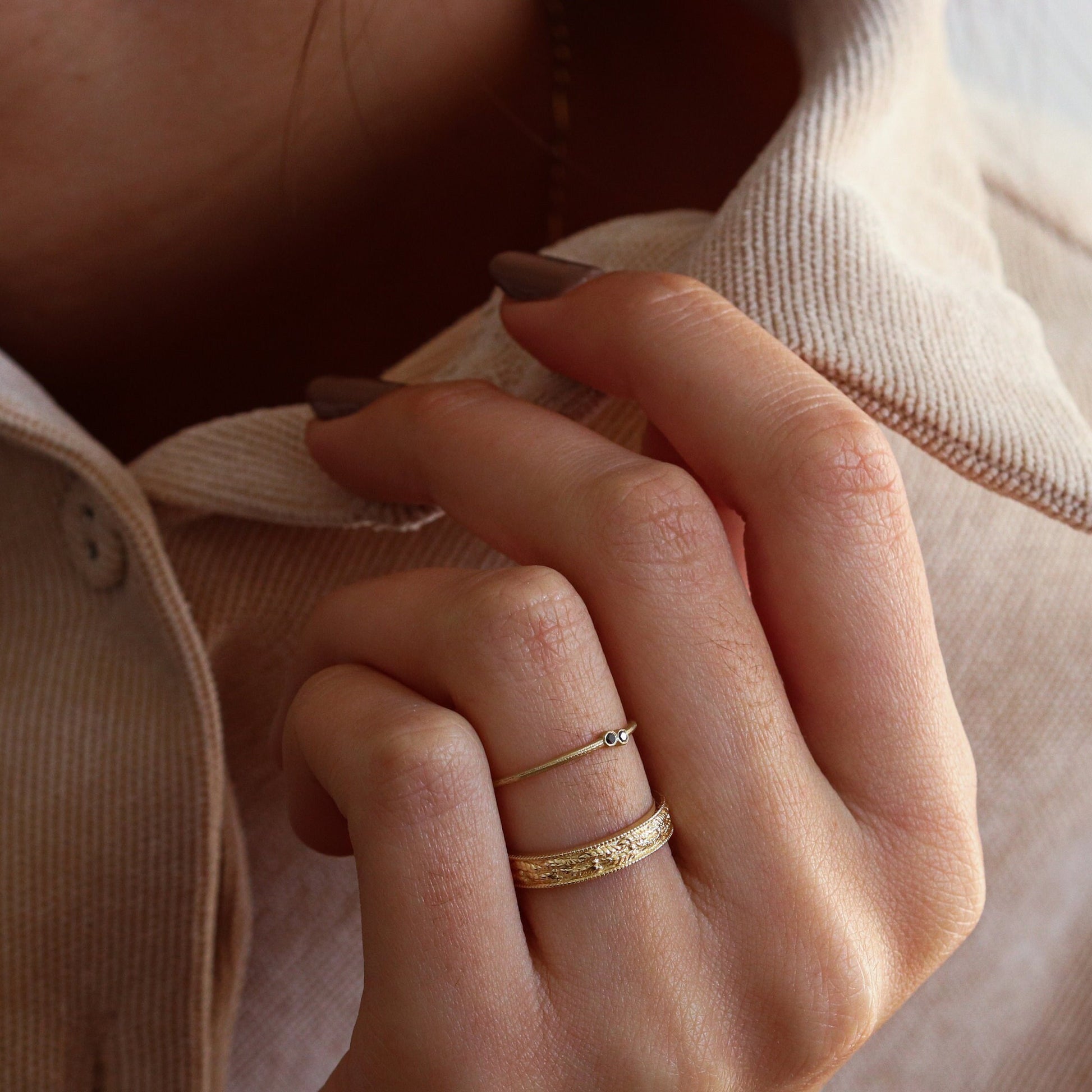 A woman's hand holding her collar, adorned with a leaf-textured and embossed gold ring, alongside one ring from the Dina Jewelry collection