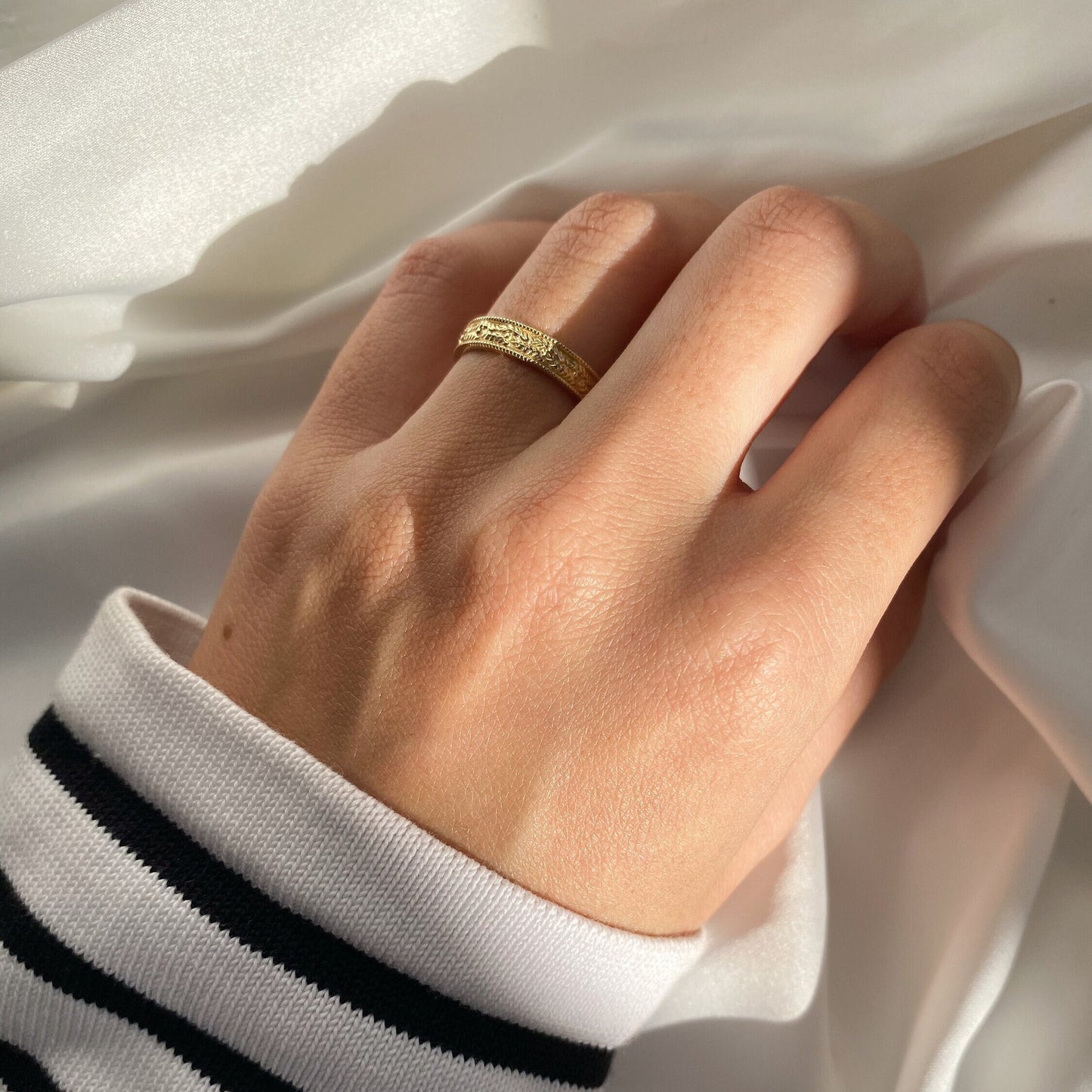 Gold ring with leaf-textured and embossed design on a woman's hand wearing a striped shirt with visible sleeve against a white background